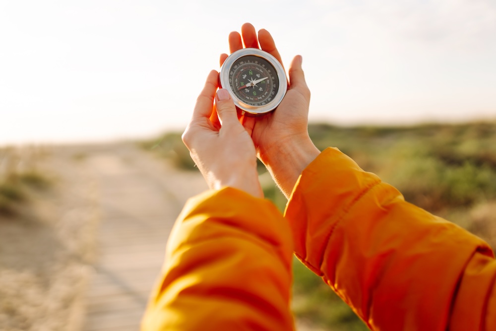 Close-up of a young female traveler's hands holding a compass against the backdrop of a sunset beach. A woman in a bright jacket looks at direction of travel and enjoys sunlight. Travel concept.